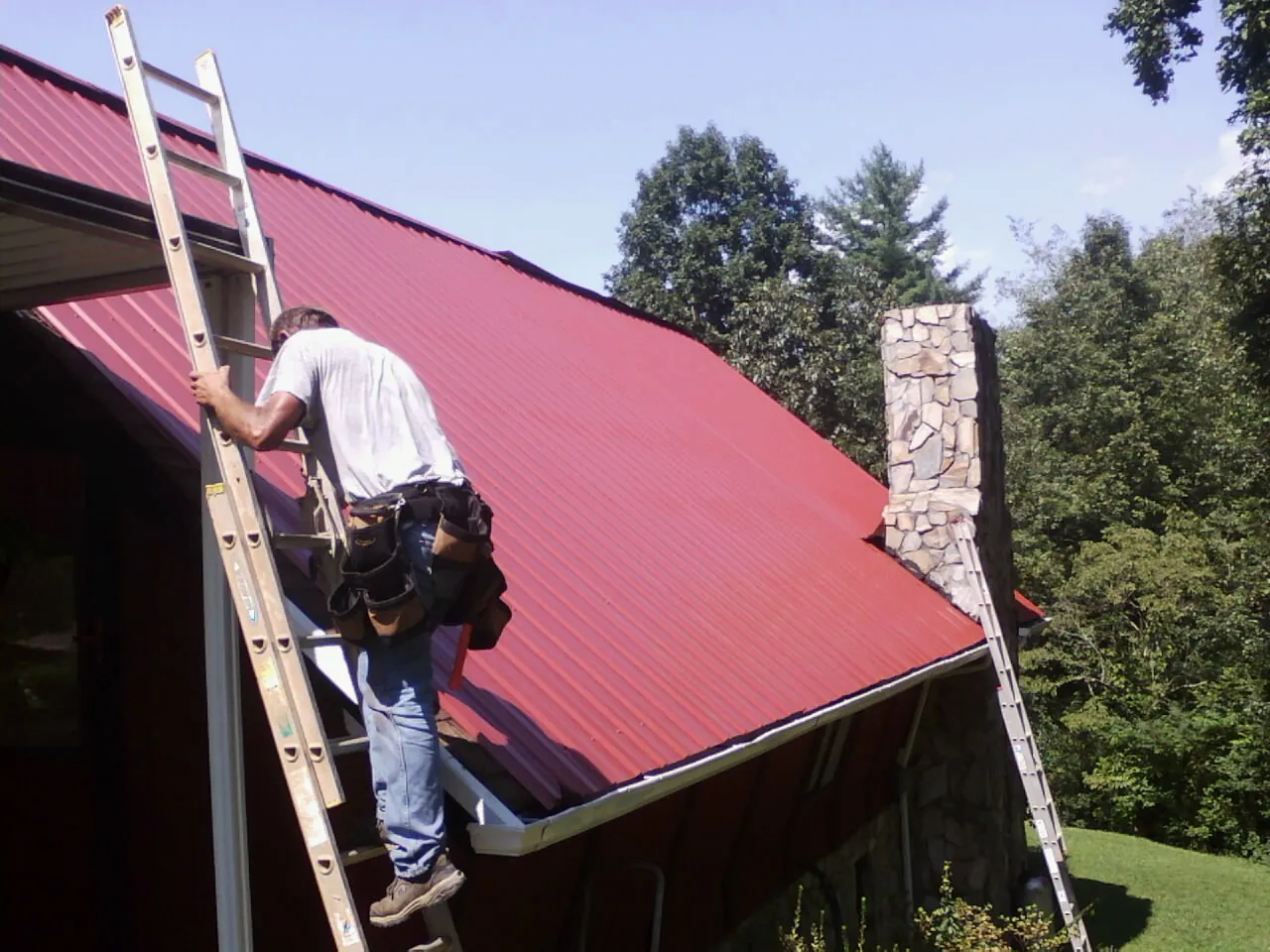 Roofer on a ladder beside a red metal roof and stone chimney — Soffit & Fascia Repair by Roofing Brothers in Mine Hill